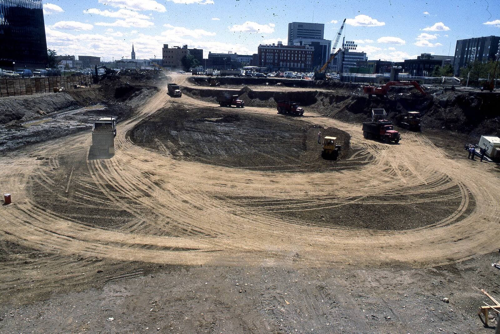 A large urban site excavated and graded, a haul road curving through it, city buildings behind.